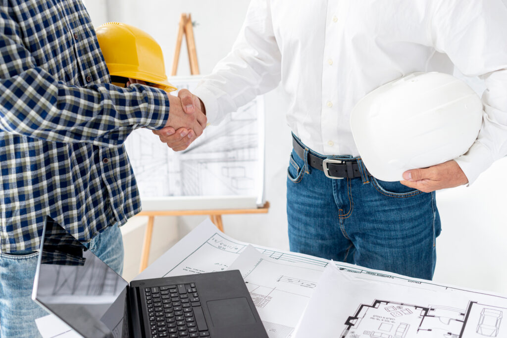 An architect and a reliable remodeling contractor shaking hands over home renovation blueprints and construction plans on a desk.