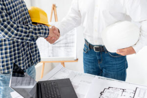 An architect and a reliable remodeling contractor shaking hands over home renovation blueprints and construction plans on a desk.